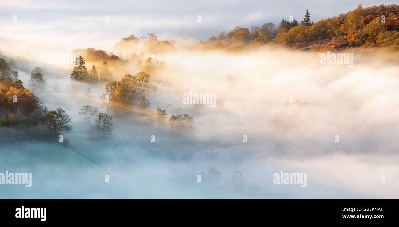 The rising sun highlights the mist floating above Loughrigg Tarn at the head of the Langdale Valley on a frosty autumn morning in the Lake District. Stock Photo