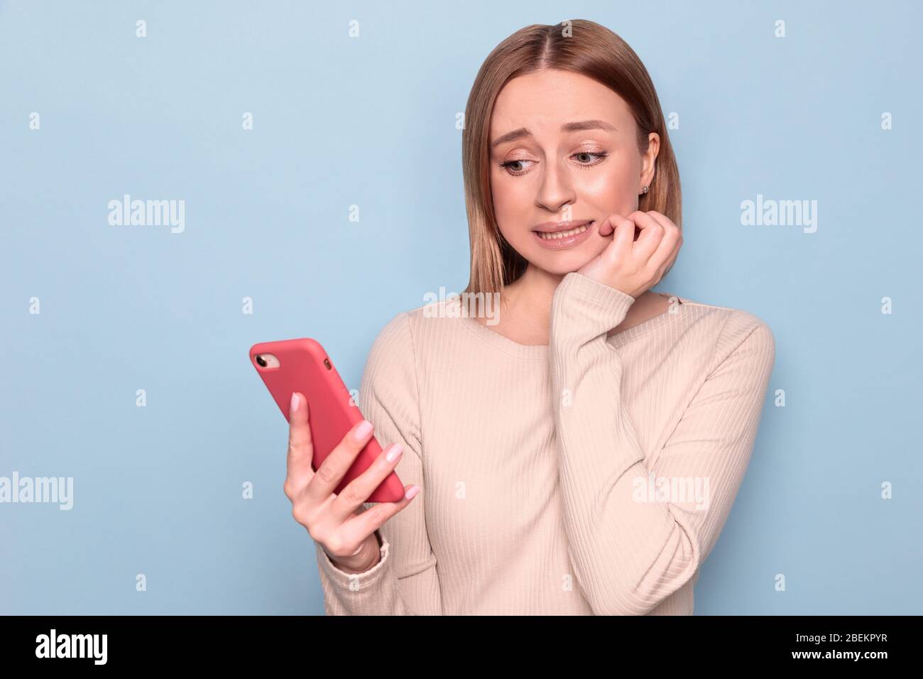 Portrait of confused nervous young woman frowning, looking at mobile phone, sent message to the wrong recipient, feeling guilty or awkward, clenching Stock Photo