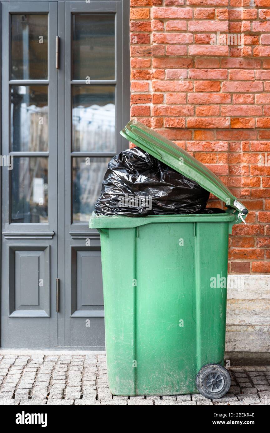 Opened big green plastic trash can with full trash bags. Disposal of waste. Overcrowded garbage can over brick wall and door. Stock Photo