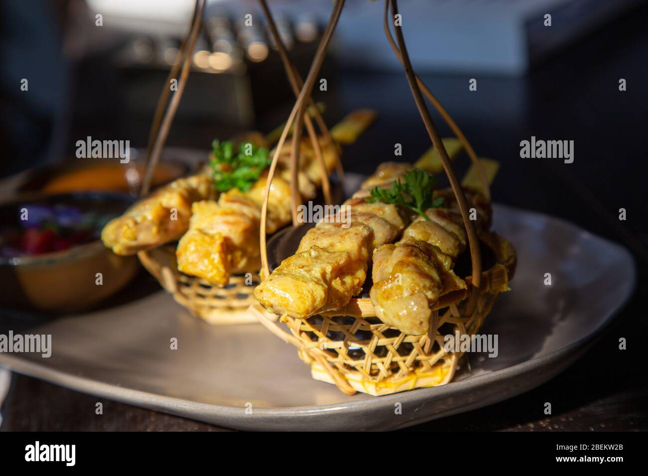 Chicken satay or Chicken kebab skewers on a black ceramic plate with side dishes made from cucumbers, red onions, peppers and Thai dipping sauce. Stock Photo