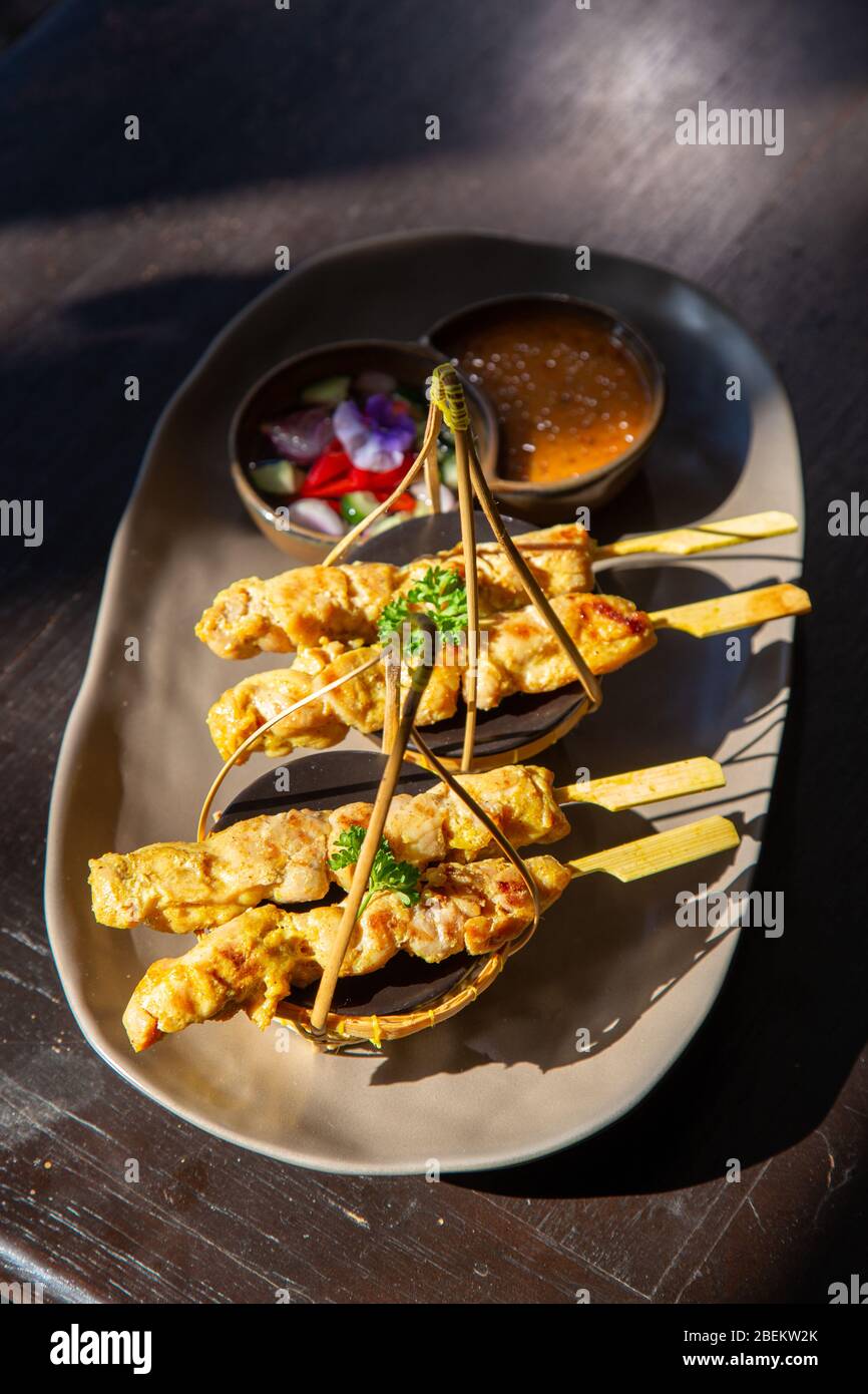 Chicken satay or Chicken kebab skewers on a black ceramic plate with side dishes made from cucumbers, red onions, peppers and Thai dipping sauce. Stock Photo