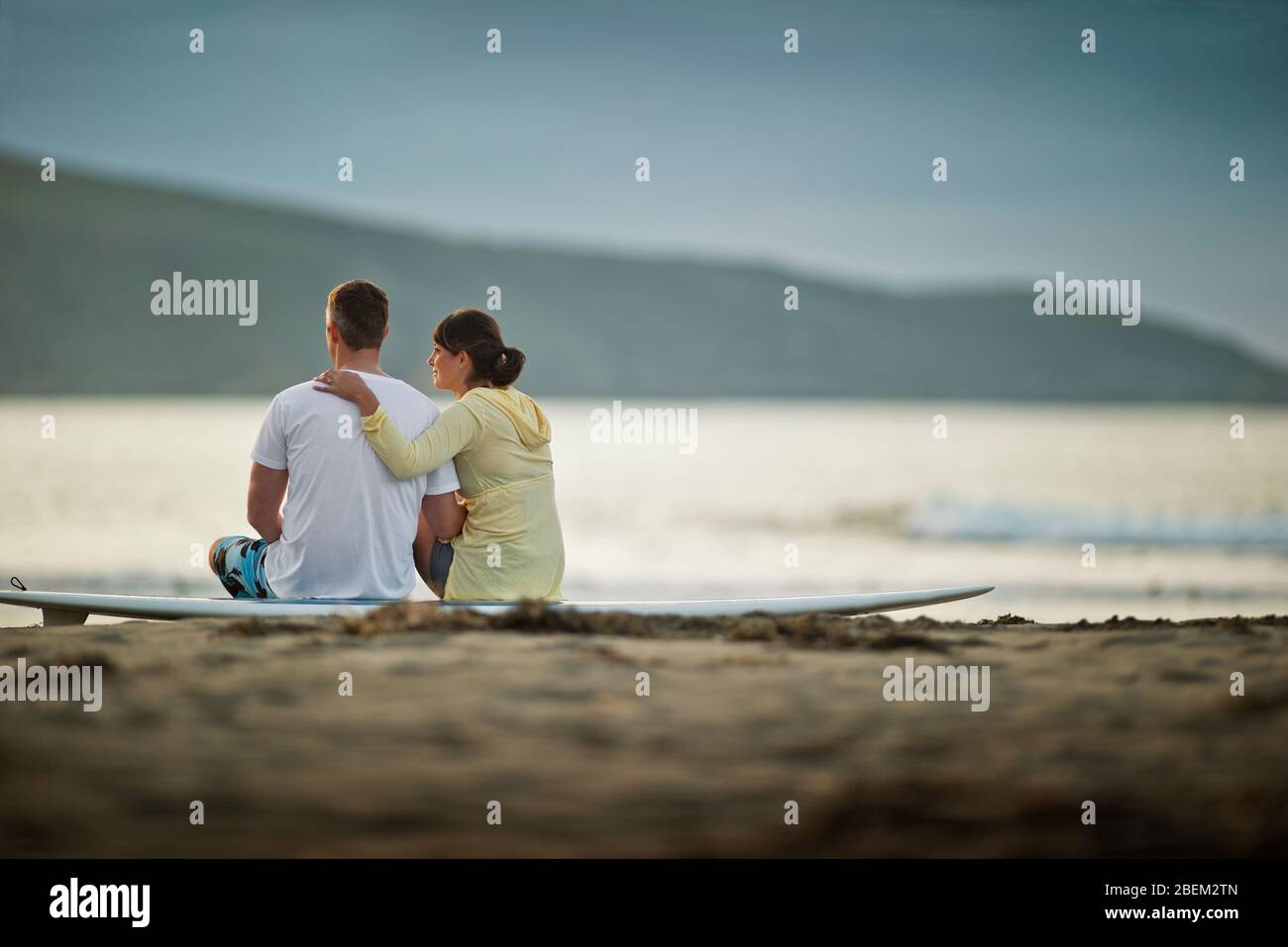 Mid adult couple sitting on a surfboard at the beach Stock Photo
