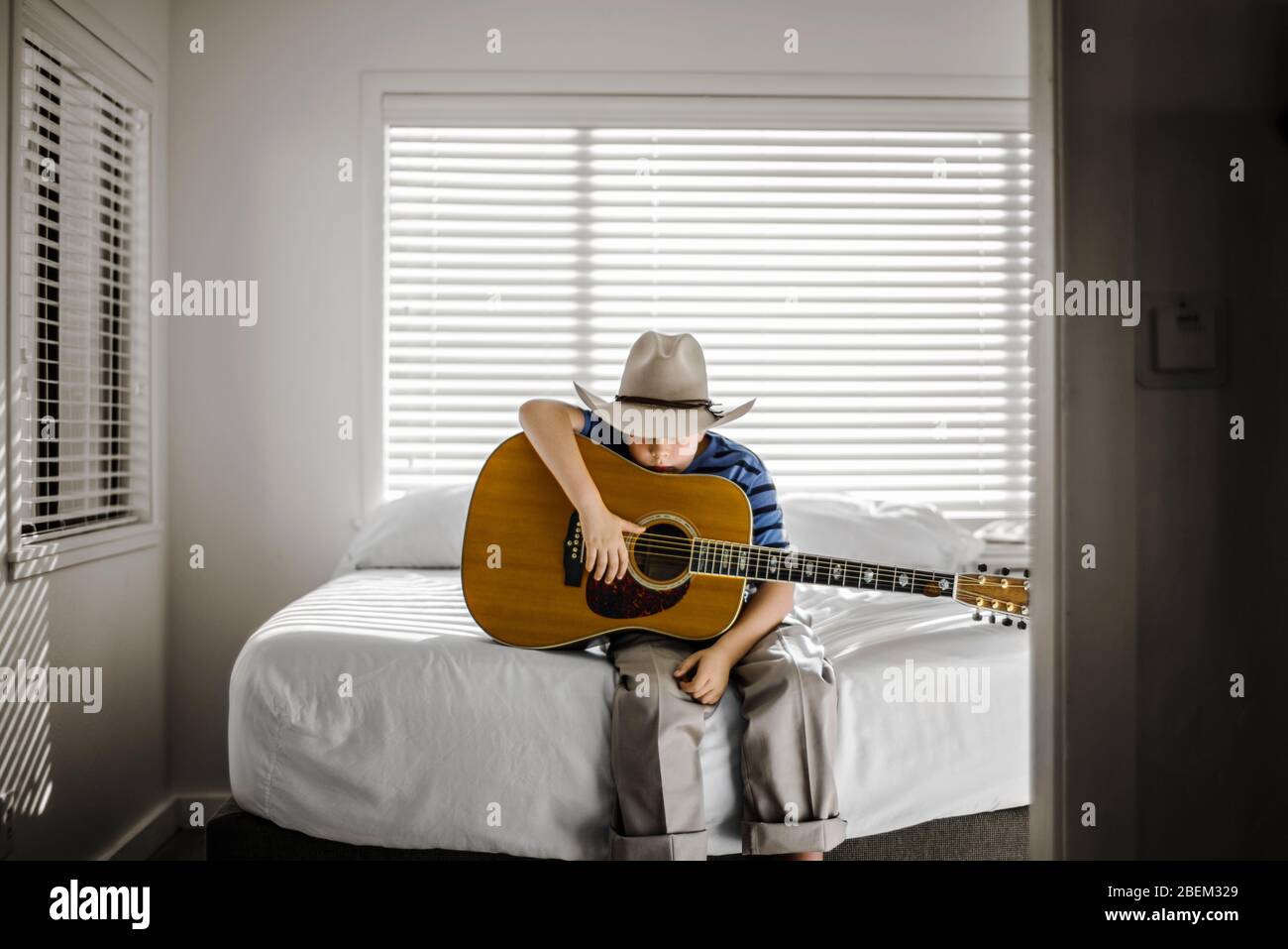 Young boy wearing a cowboy hat sitting on his bed holding an acoustic guitar Stock Photo