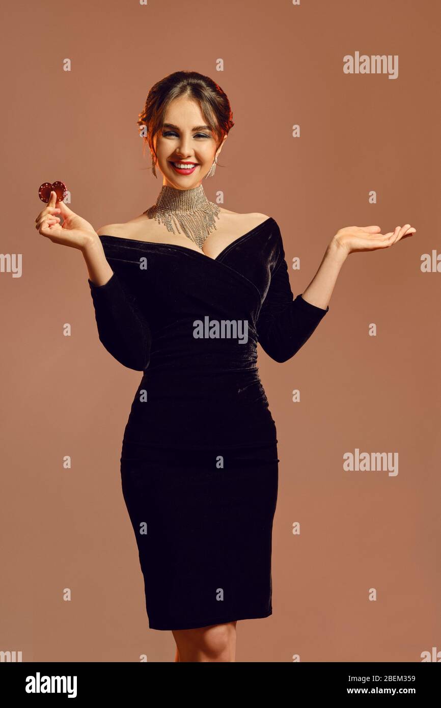 Brunette girl in black dress, jewelry. Smiling, showing two red chips and  holding something, posing on brown background. Poker, casino. Close-up  Stock Photo - Alamy