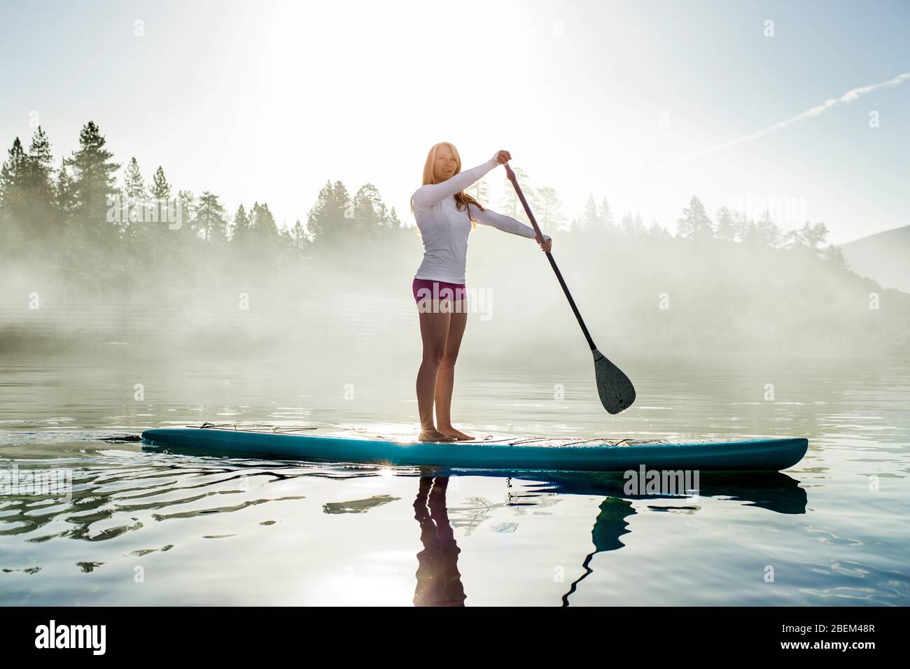 Active young woman paddle boarding across a lake in the early morning fog Stock Photo