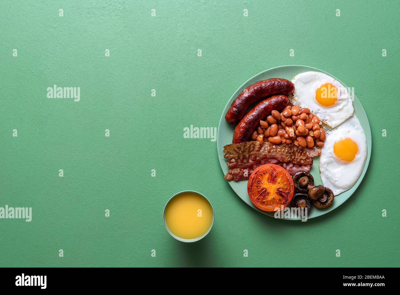 Traditional english breakfast with baked beans, sausages, bacon and sunny-side-up eggs on a green background. British full breakfast on a plate. Stock Photo