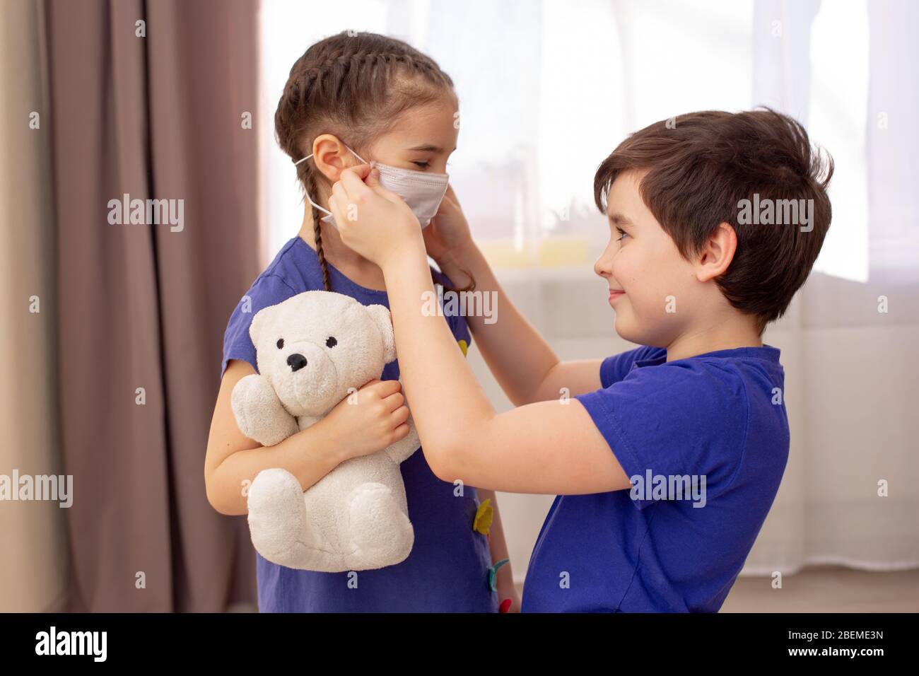 A boy with a sweet smile in a blue T-shirt is standing in the room, putting on a white medical mask to his little sister in a blue dress and a white t Stock Photo