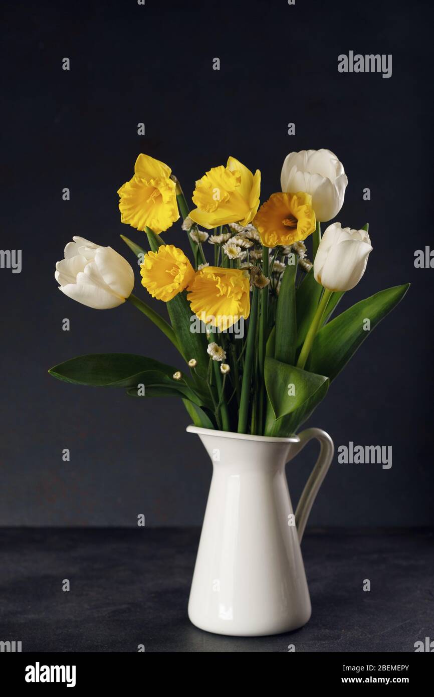 Fresh bouquet of yellow daffodils and white tulips in vase on black table. Springtime still life concept. Stock Photo
