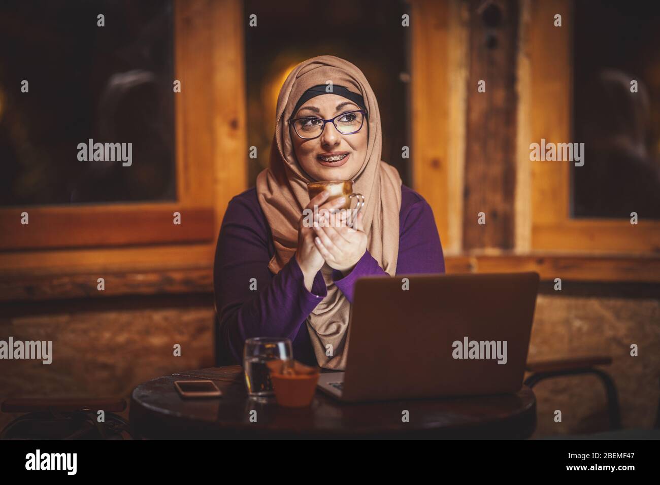 Middle aged Muslim woman wearing hijab is sitting on the cafe in urban environment, drinking coffee and working on laptop. Stock Photo