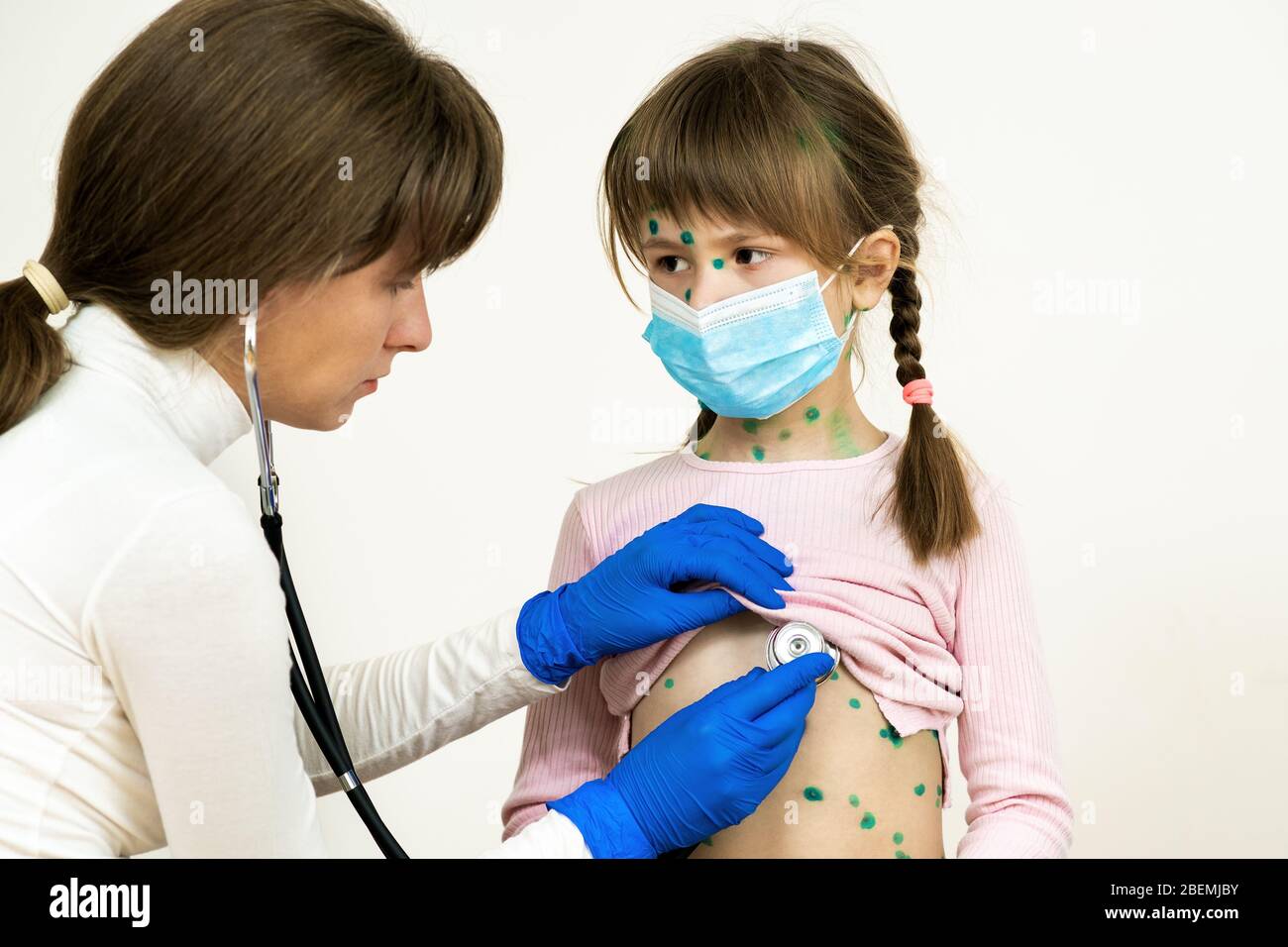 Doctor examining child girl covered with green rashes on face and stomach ill with chickenpox, measles or rubella virus. Stock Photo