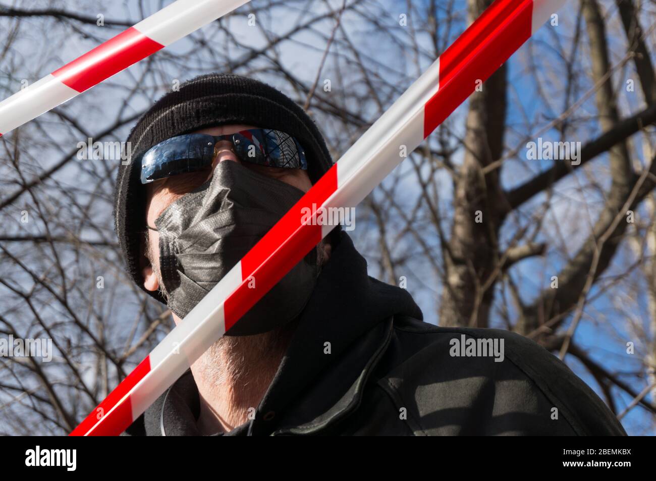 A man in a black protective mask, hat and sunglasses looks at the sky in front of a restrictive red and white ribbon. A concept quarantine during the Stock Photo