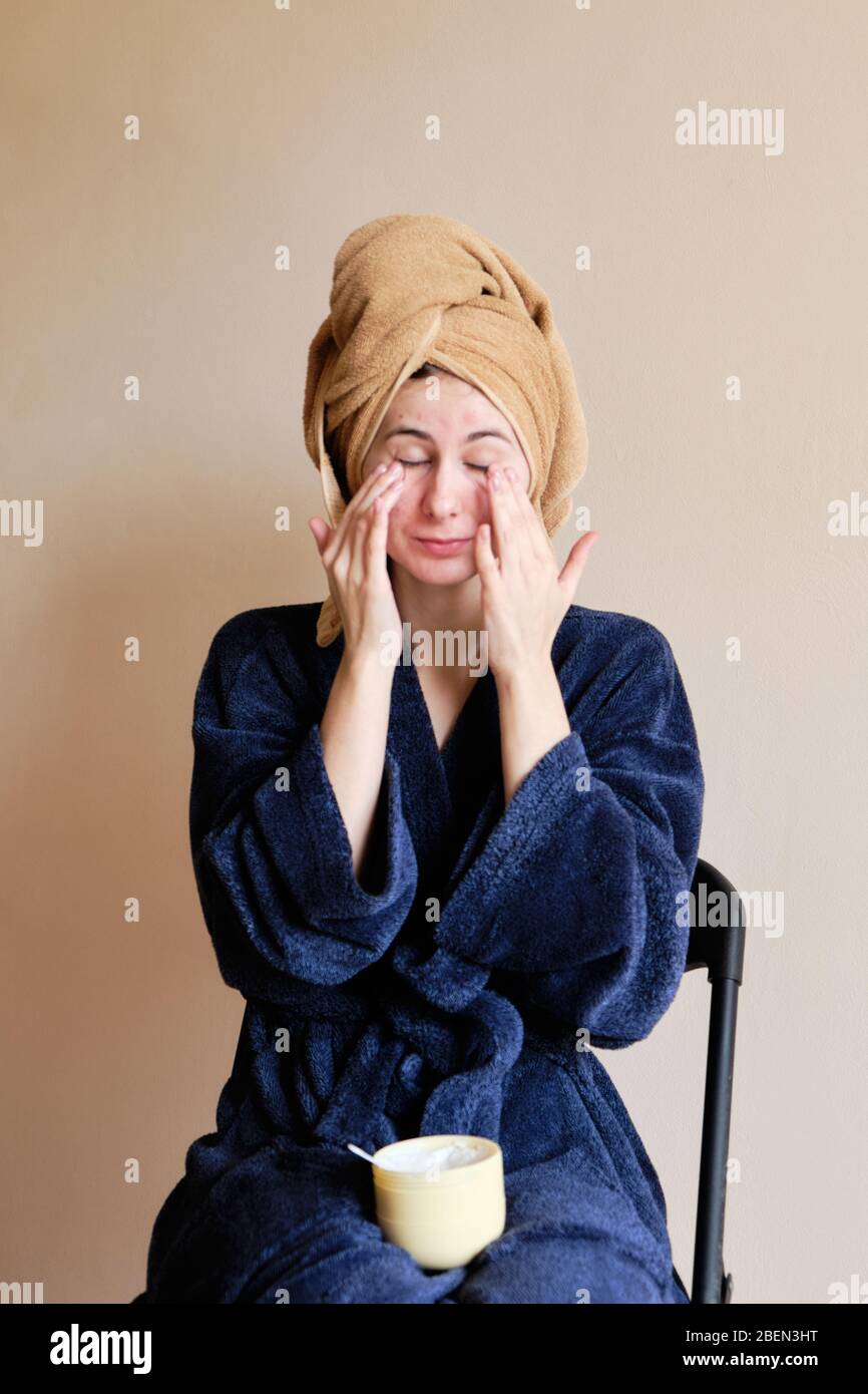 woman in a robe and with a towel on her head applies cosmetics to skin Stock Photo