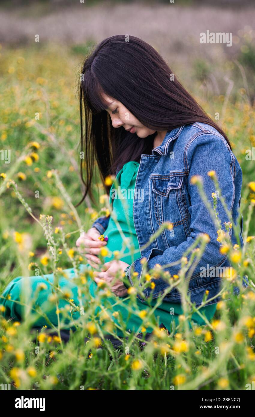 Beautiful pregnant young Asian woman with long black hair wearing a green dress and blue denim jacket holding her belly as she sits in a green field Stock Photo