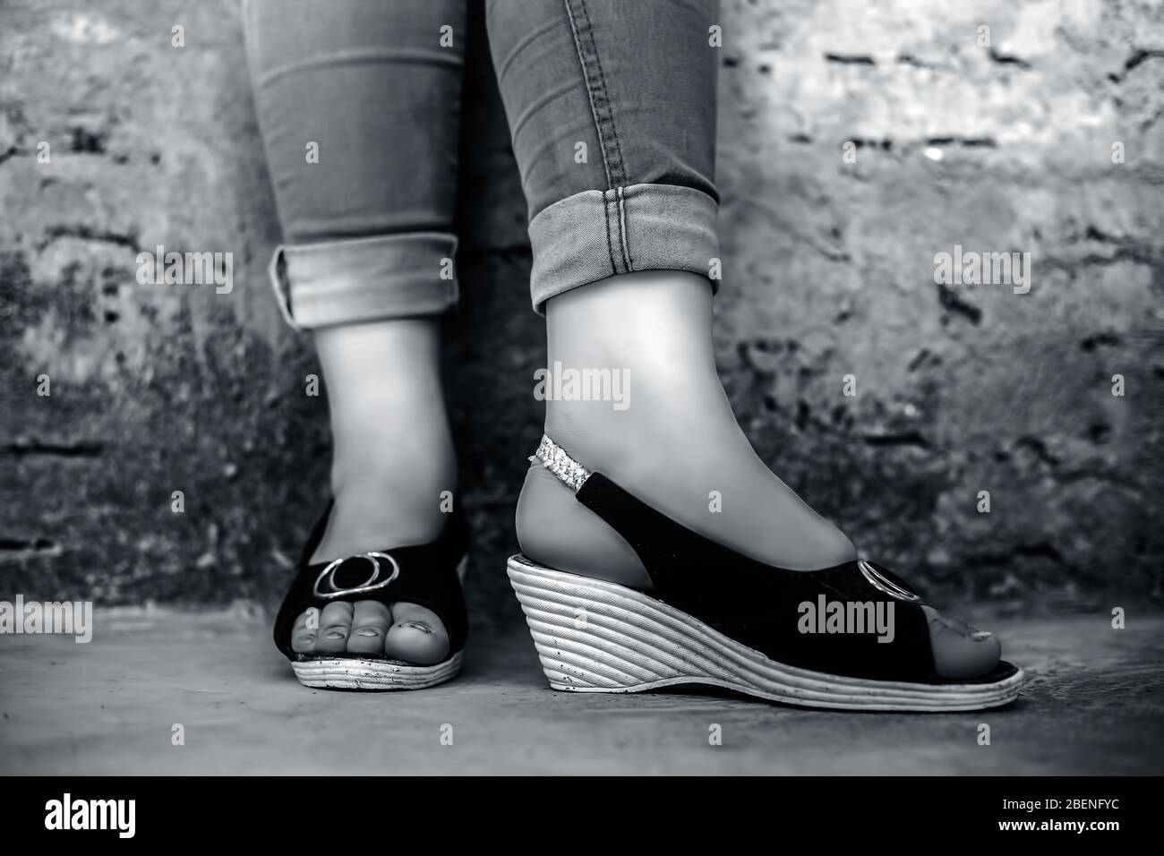 Shot of foot of elegant young teen girl wearing blue denim jeans and peep-toe sandals posing crossed legs against brick wall background in black and w Stock Photo