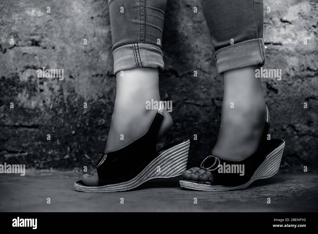 Shot of foot of elegant young teen girl wearing blue denim jeans and peep-toe sandals posing crossed legs against brick wall background in black and w Stock Photo