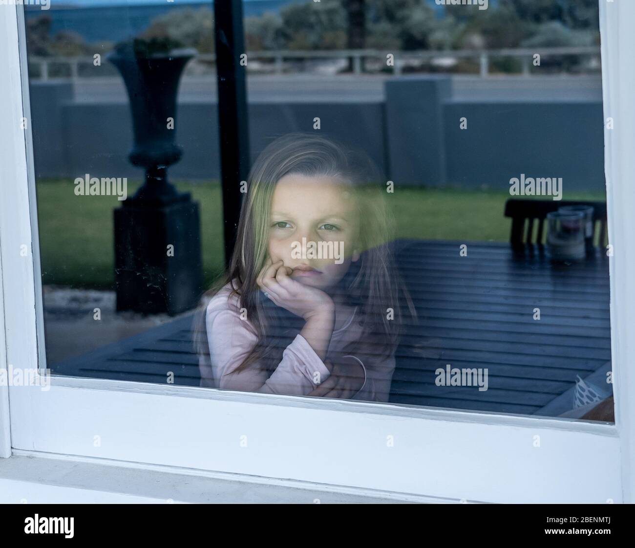COVID-19 Lockdowns. Depressed and lonely little girl looking through the window during quarantine. Child feeling sad as coronavirusu pandemic forces f Stock Photo