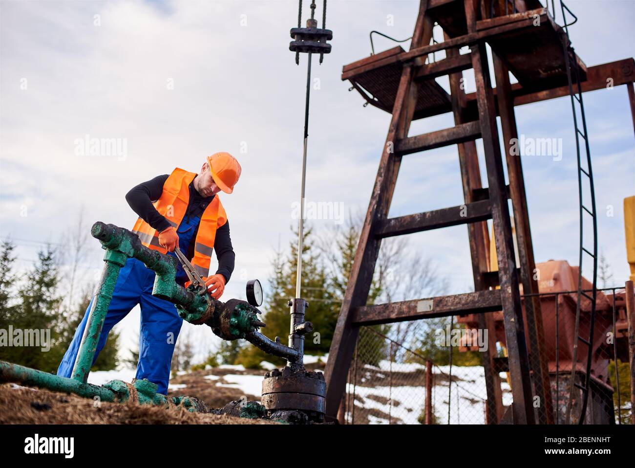 Oil worker in blue overalls, orange vest and helmet working with wrenching a bolt on a gas pipe near an oil pump jack, concept of oil industry Stock Photo