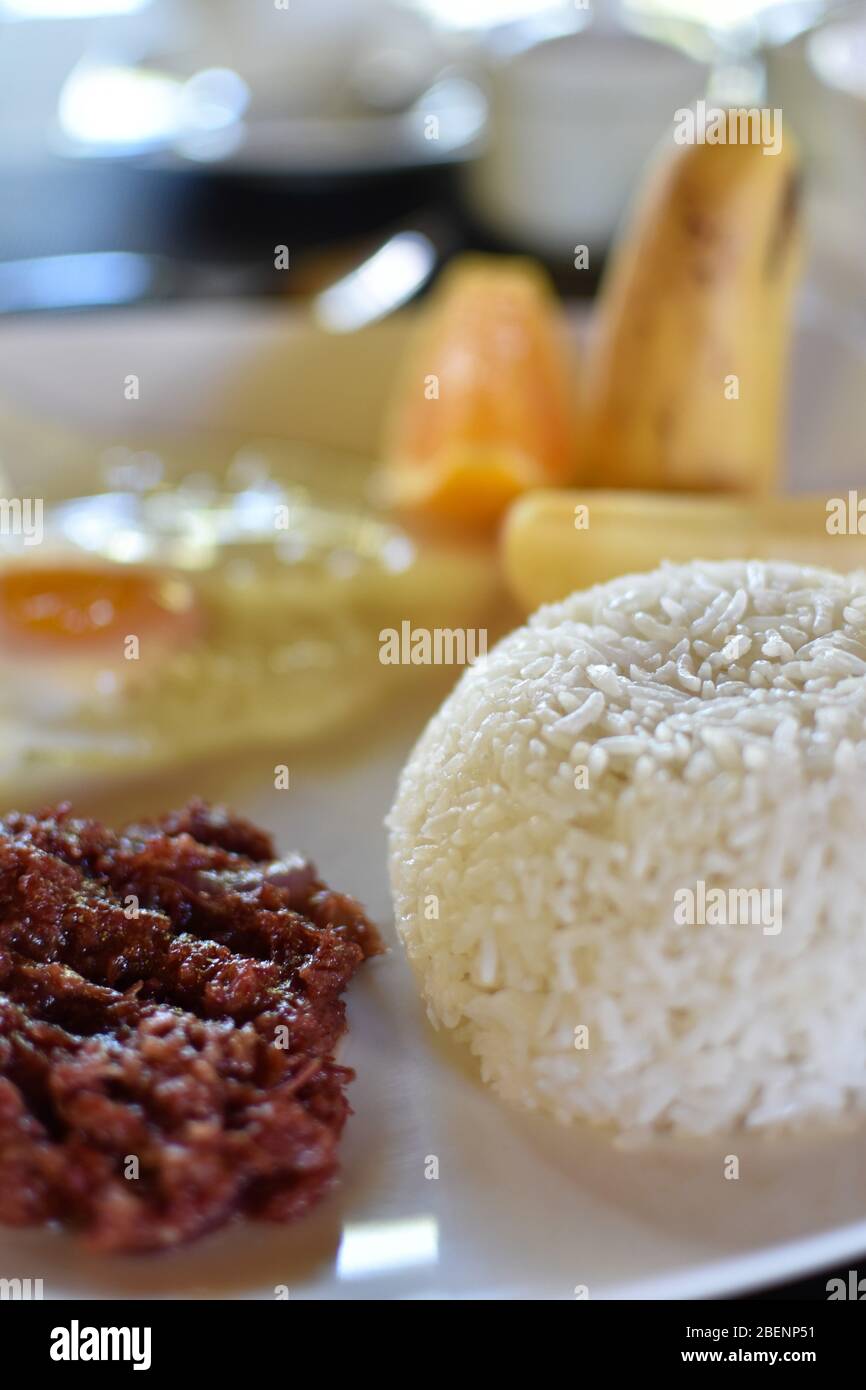 A plate of breakfast consisting of white rice, corned beef, fried egg, and sliced fruits Stock