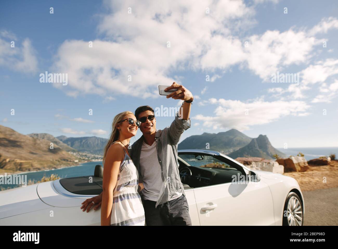 Man taking selfie with his girlfriend on a road trip. Couple standing by their car taking selfie with a mobile phone outdoors. Stock Photo