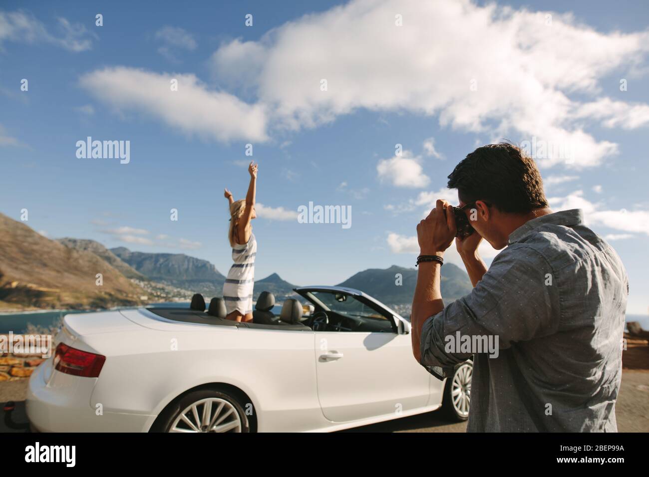 Man taking pictures of a woman standing in a convertible car. Couple taking pictures on road trip. Stock Photo