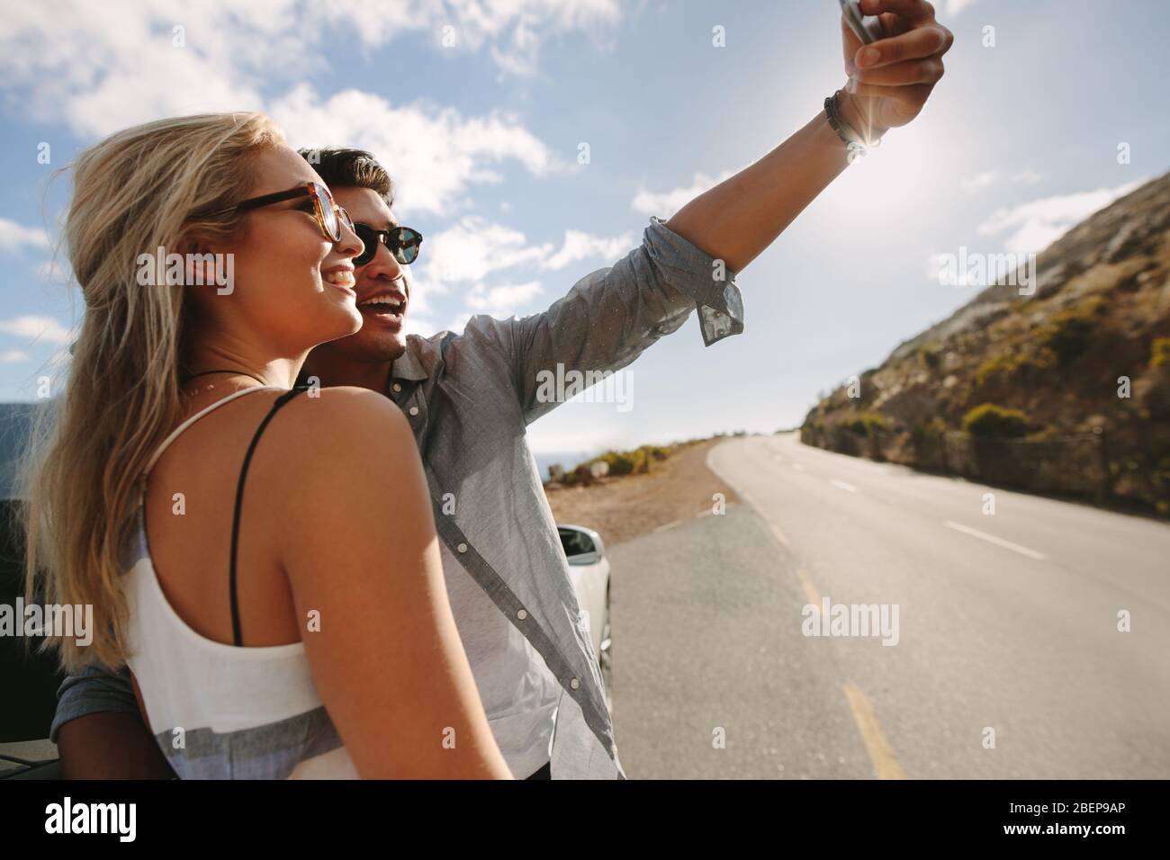 Couple taking a selfie using a smart phone and smiling while standing near car during travel. Man and woman posing for a self portrait by their car wh Stock Photo