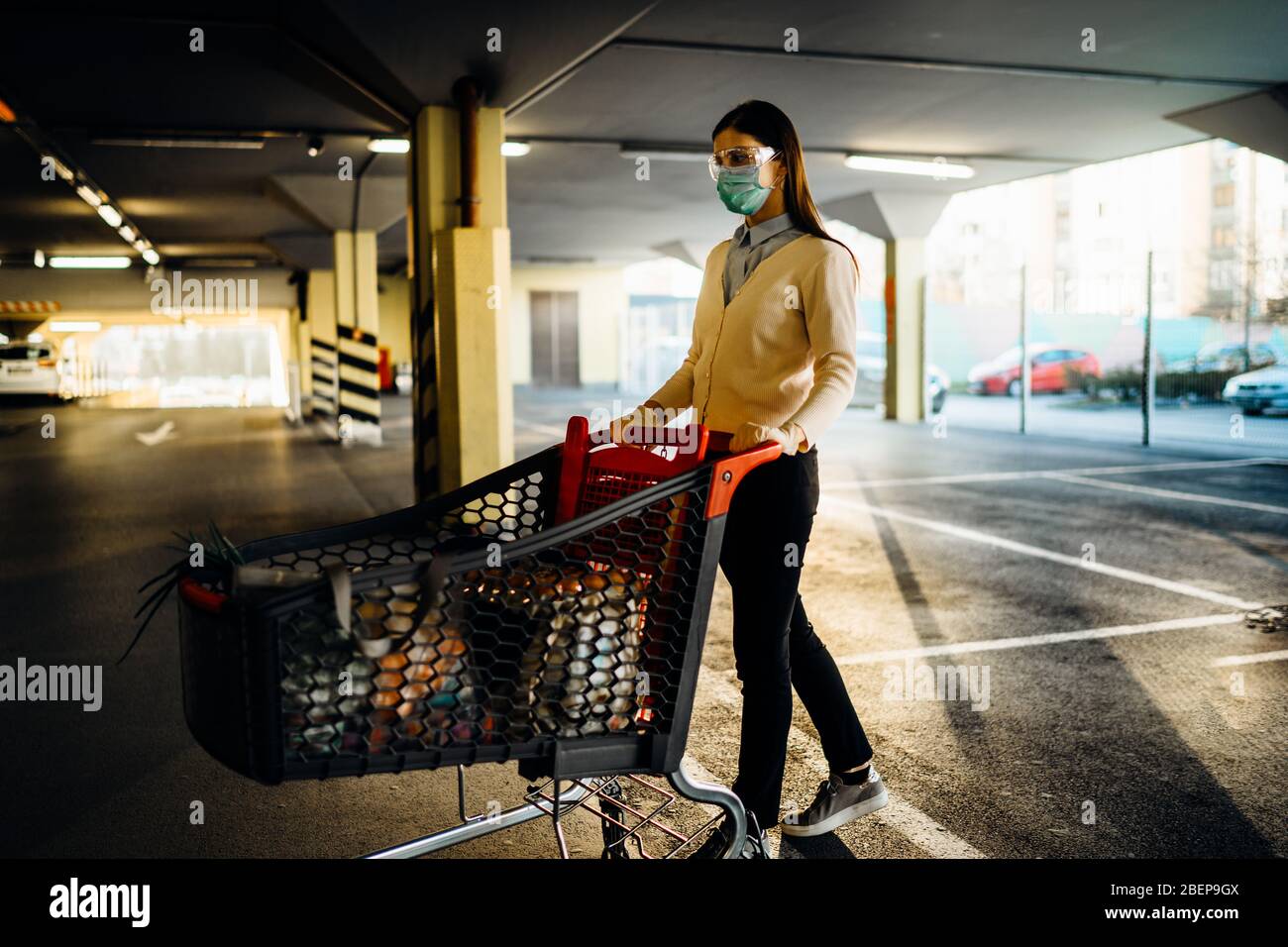 Woman wearing mask groceries/supplies shopping in supermarket,pushing trolley.Food supplies shortage.Panic buying and hoarding.Empty parking garage.Su Stock Photo