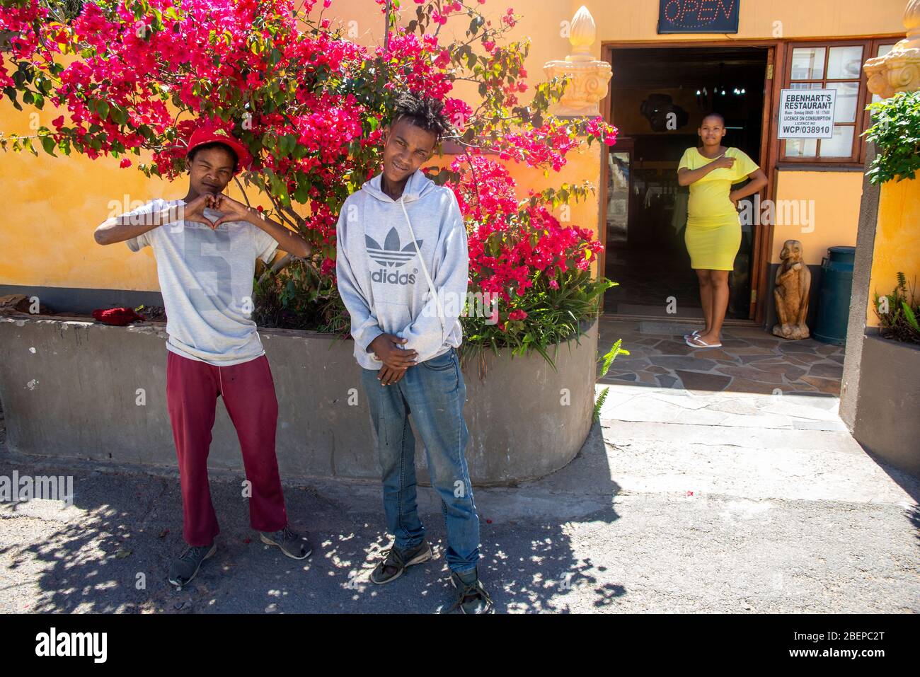 Two teenage street boys pose in front of red Bougainvillea on the side of the road. A waitress stands in the door of a cafe behind them. Their plain colours contrast with the strong colours of the wall and flowers.  One of them makes a gang sign. They are probably brothers and they live in Oudtshoorn in the Western Cape. Stock Photo