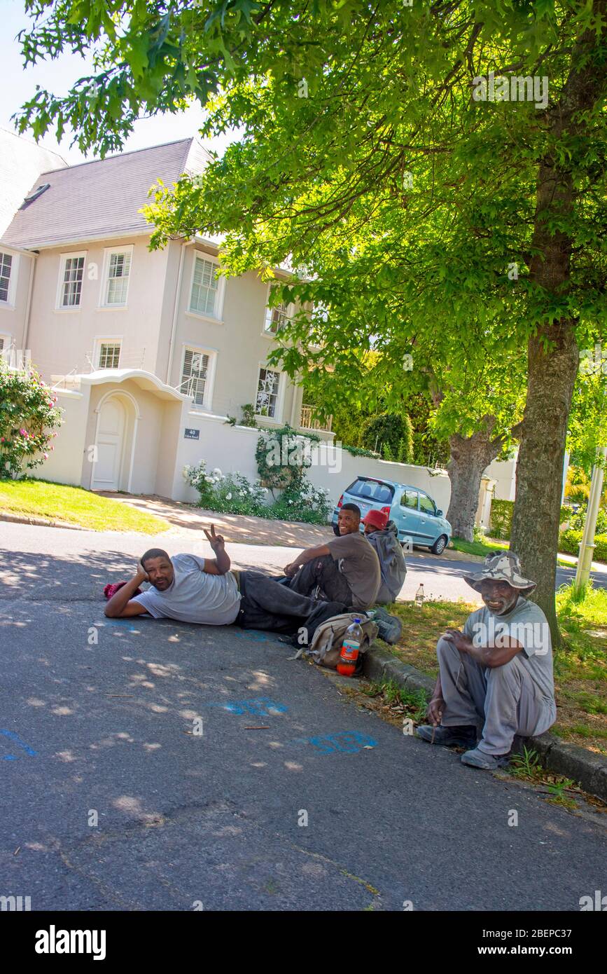 Black building workers rest on the roadside during the day. They are working in an exclusive and upmarket area of Cape Town called Claremont. The men are friendly and happy. There is a nice mansion behind them and a beautiful tree. Both bask in the sunshine. The residents are mostly white and they create a lot of work for the community. Stock Photo
