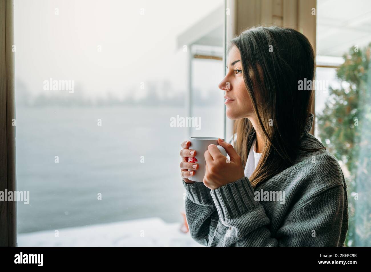 Young woman spending free time home.Self care,staying home.Enjoying view,gazing through to the window.Quarantined person indoors.Serene mornings. Stock Photo