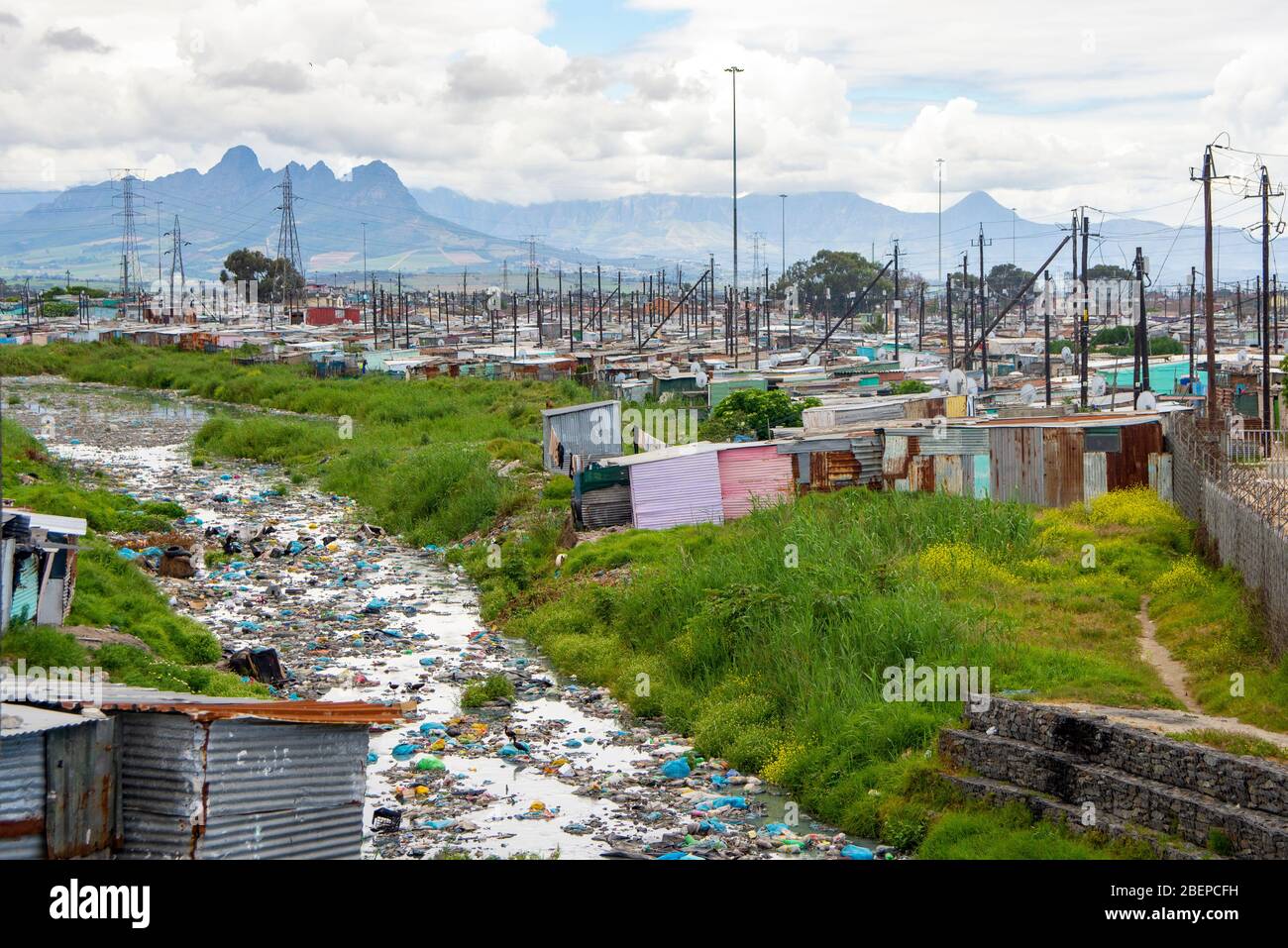 A river is full of plastic and other rubbish as it runs through a township in Cape Town. The polluted river is in the Khayelitsha slum. The area contrasts with the lush countryside in the background. No effort appears  to be made to clear the river. Stock Photo