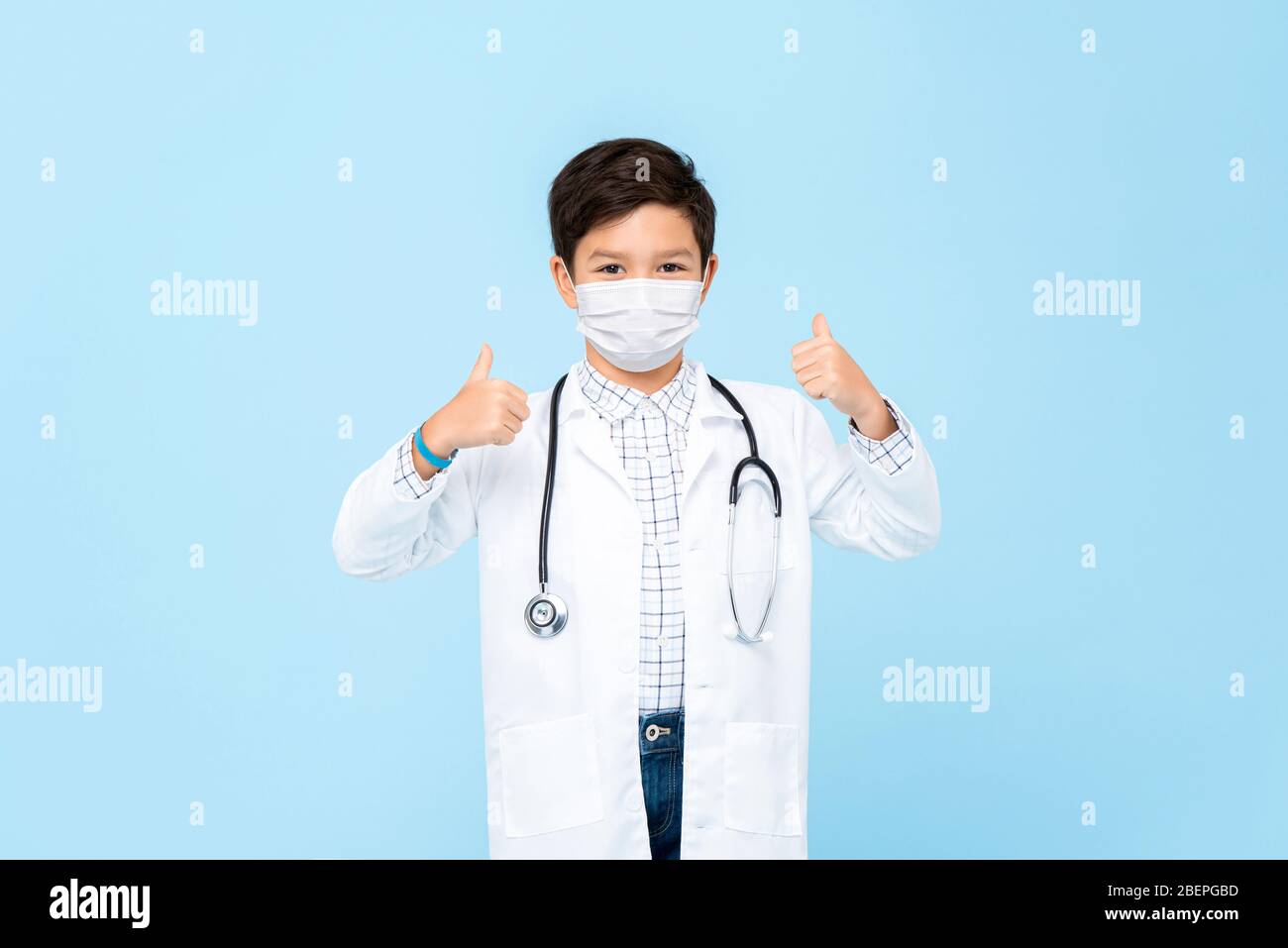 Cute boy doctor wearing medical mask and giving thumbs up isolated on light blue background Stock Photo