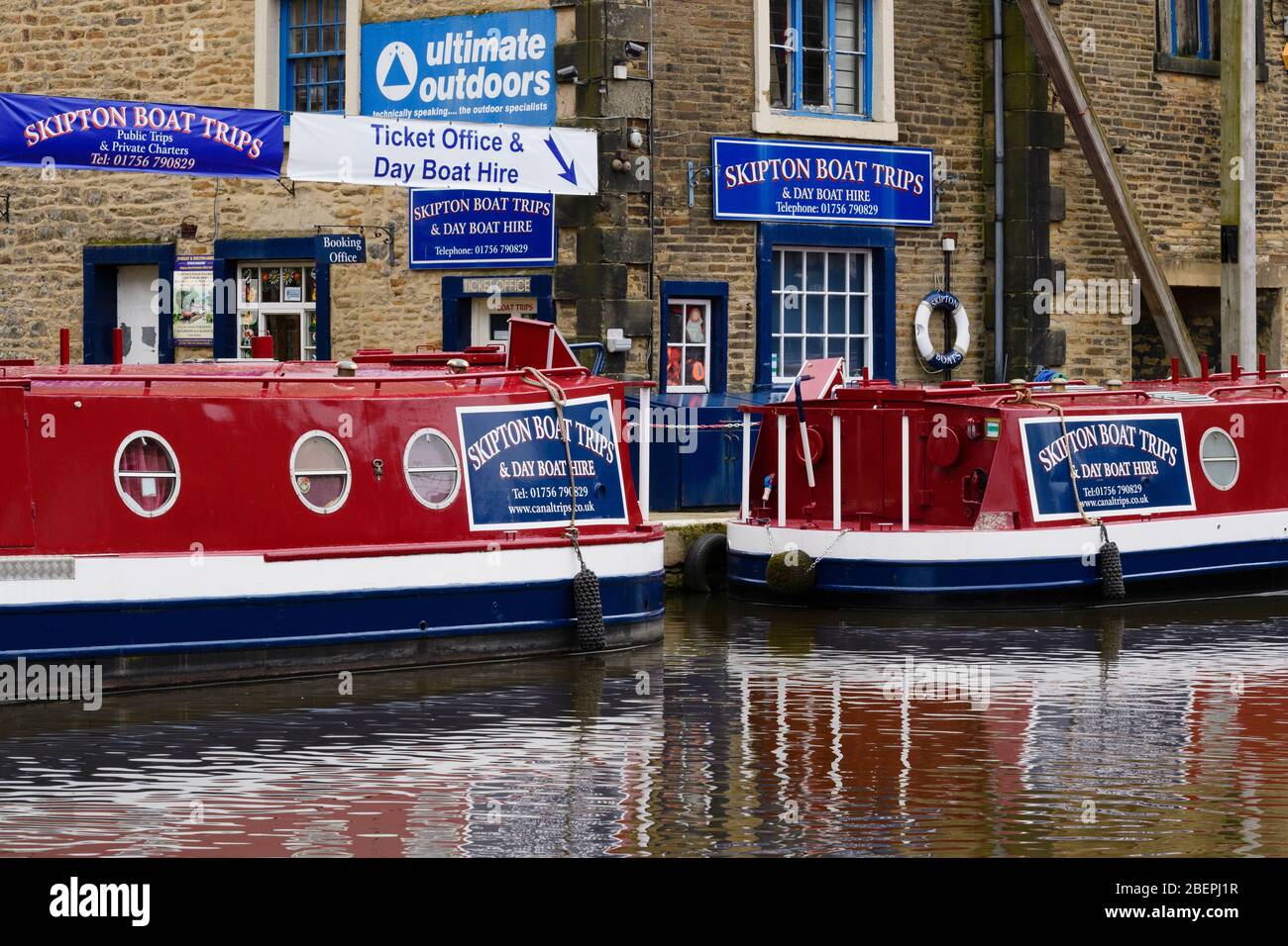 2 bright red blue canal narrow boats moored on water by boat hire business ticket office & banner - Leeds-Liverpool Canal, Skipton, Yorkshire, England Stock Photo
