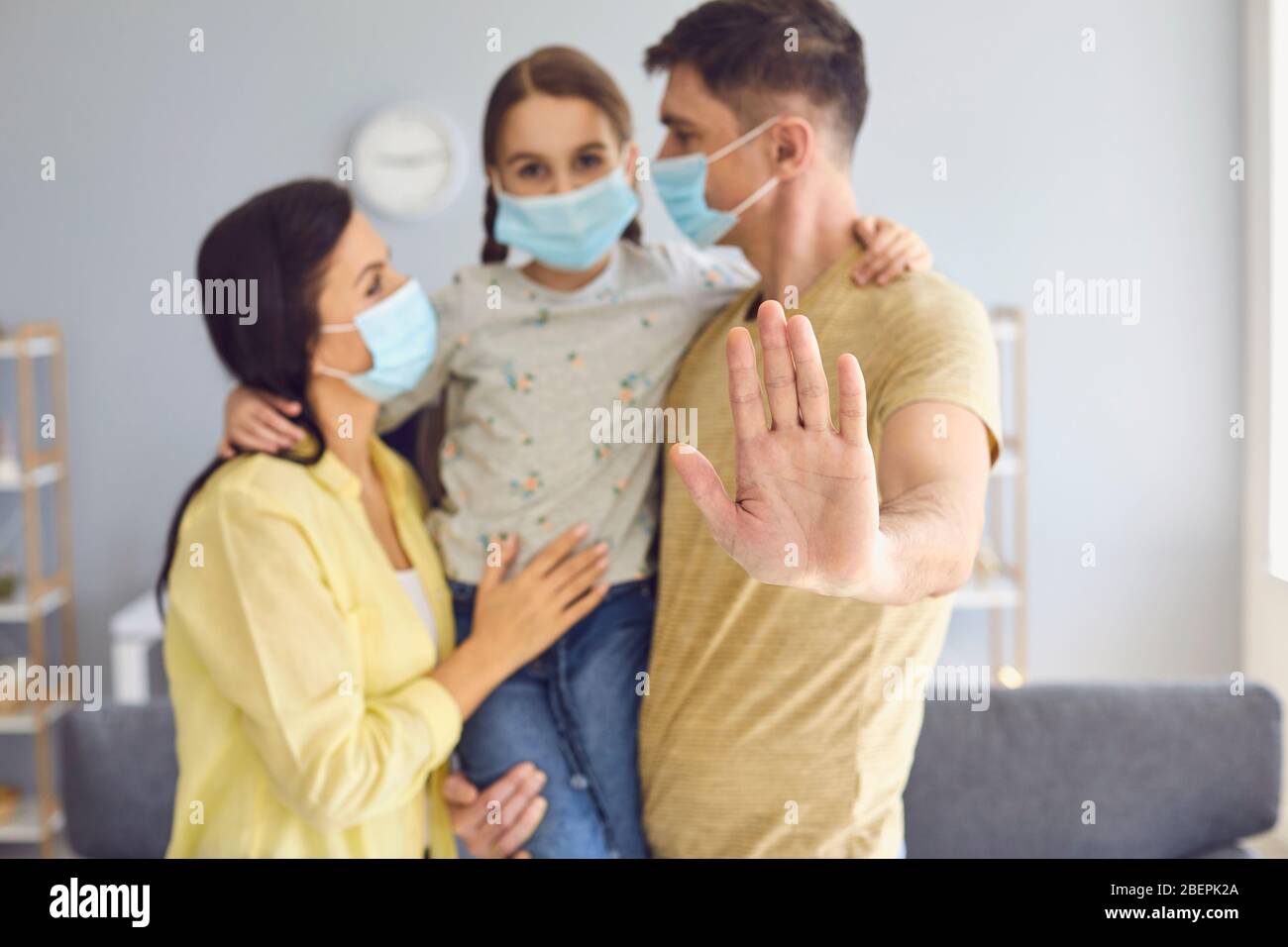 Family in medical masks on their faces looks at the camera while standing in a room at home. Father hand forward up concept of protecting a family of Stock Photo