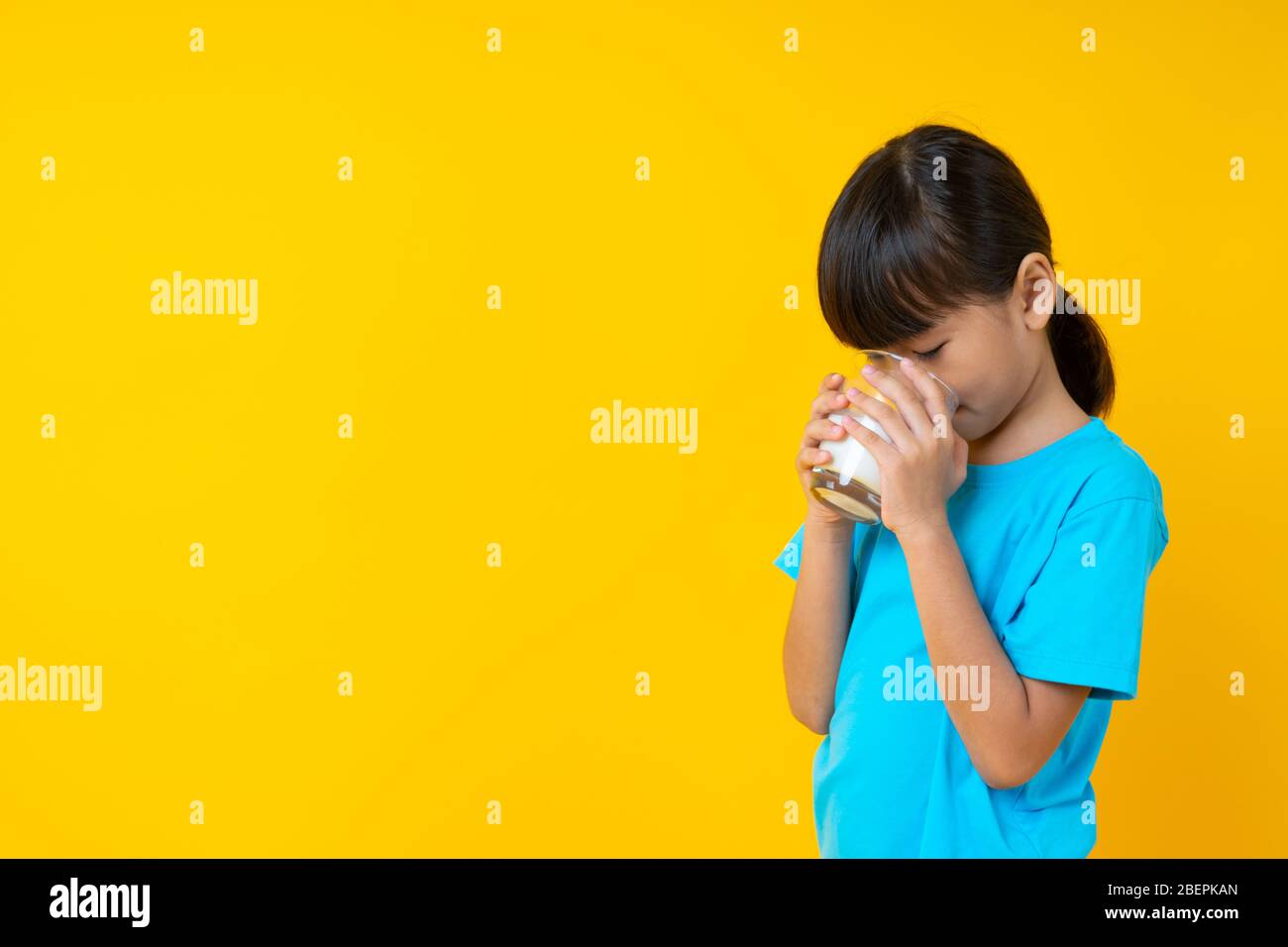 Happy Thai kid holding glass of milk isolated, young Asian girl drinking milk for strong health on yellow background Stock Photo