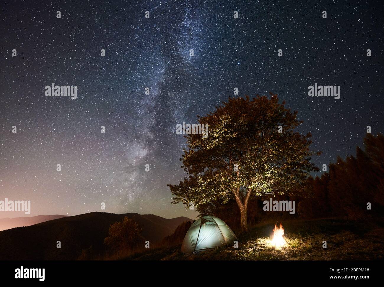 Tourist Camping Near Forest At Night Illuminated Tent And Bonfire Under Magical Night Sky Full Of Stars And Milky Way On The Background Incredibly Beautiful Starry Sky And Mountains Stock Photo