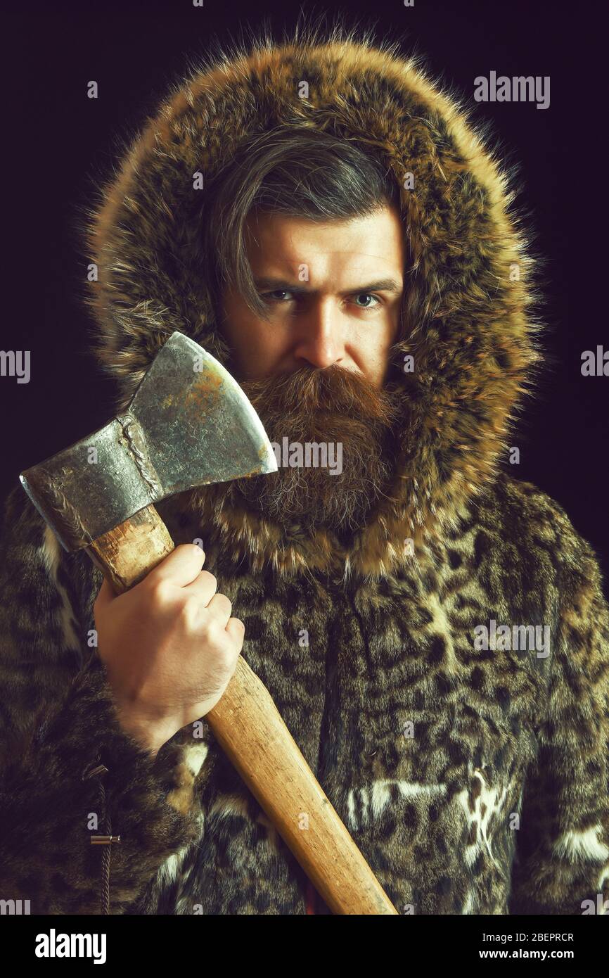 handsome bearded man or woodman guy with fashionable mustache and beard on serious face in brown fur coat, holds sharp axe, or ax on black background Stock Photo