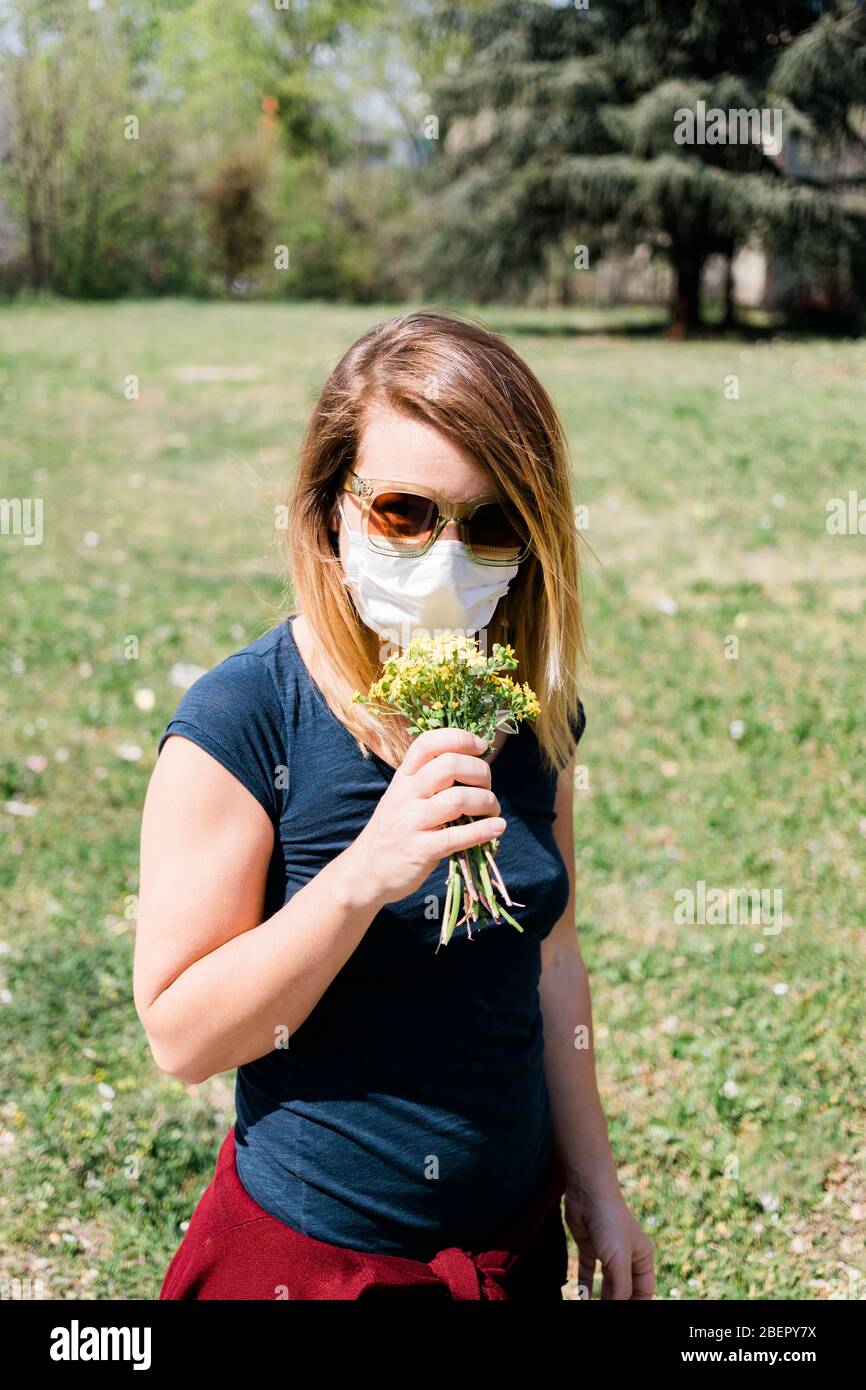 Young woman wearing surgical mask trying to smell wild flowers Stock Photo