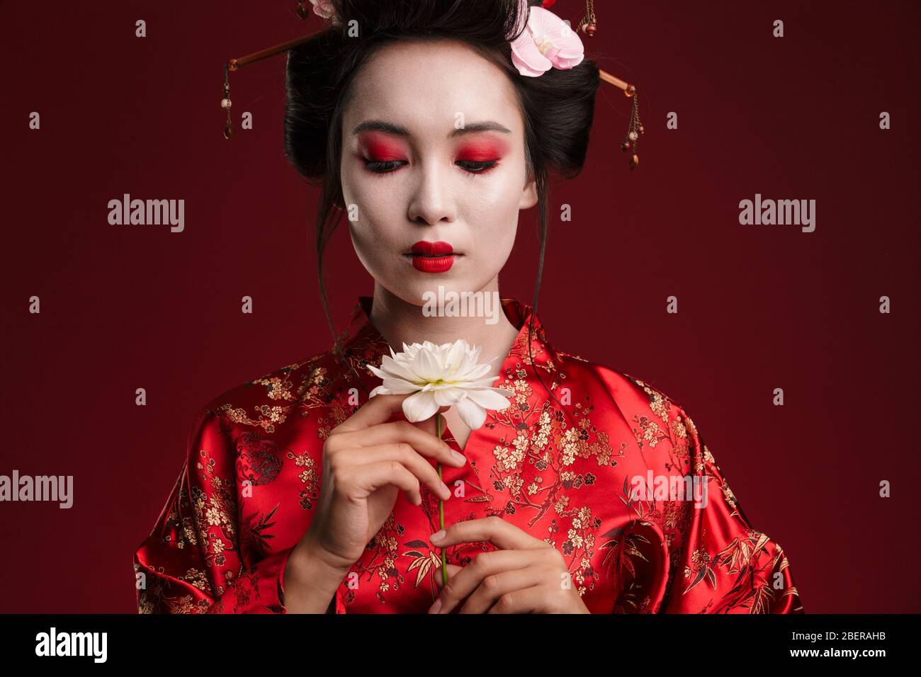 Image of beautiful geisha woman in traditional japanese kimono holding flower isolated over red background Stock Photo