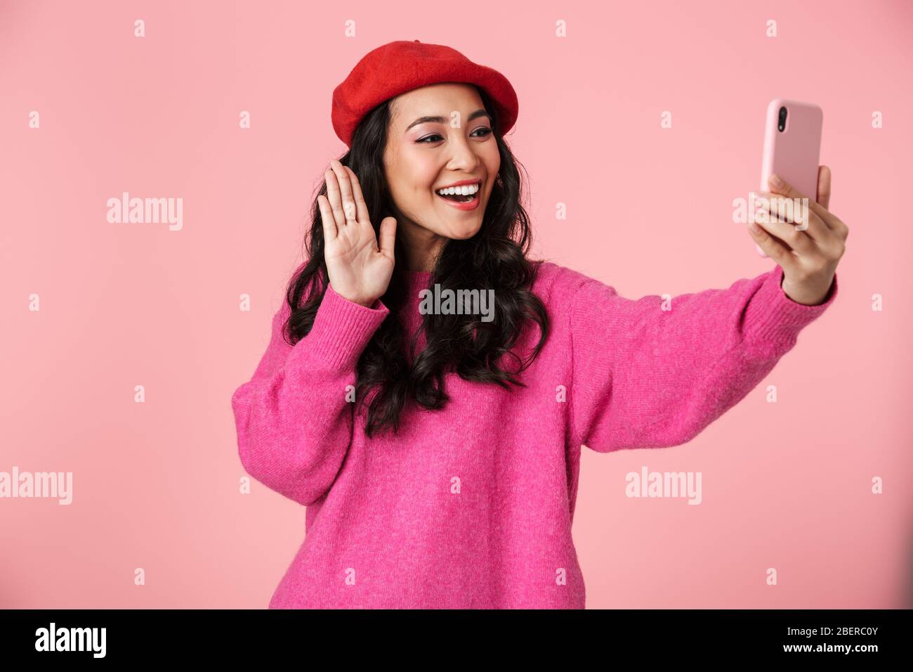 Image of joyful beautiful asian girl with long dark hair wearing beret smiling and waving at smartphone isolated over pink background Stock Photo