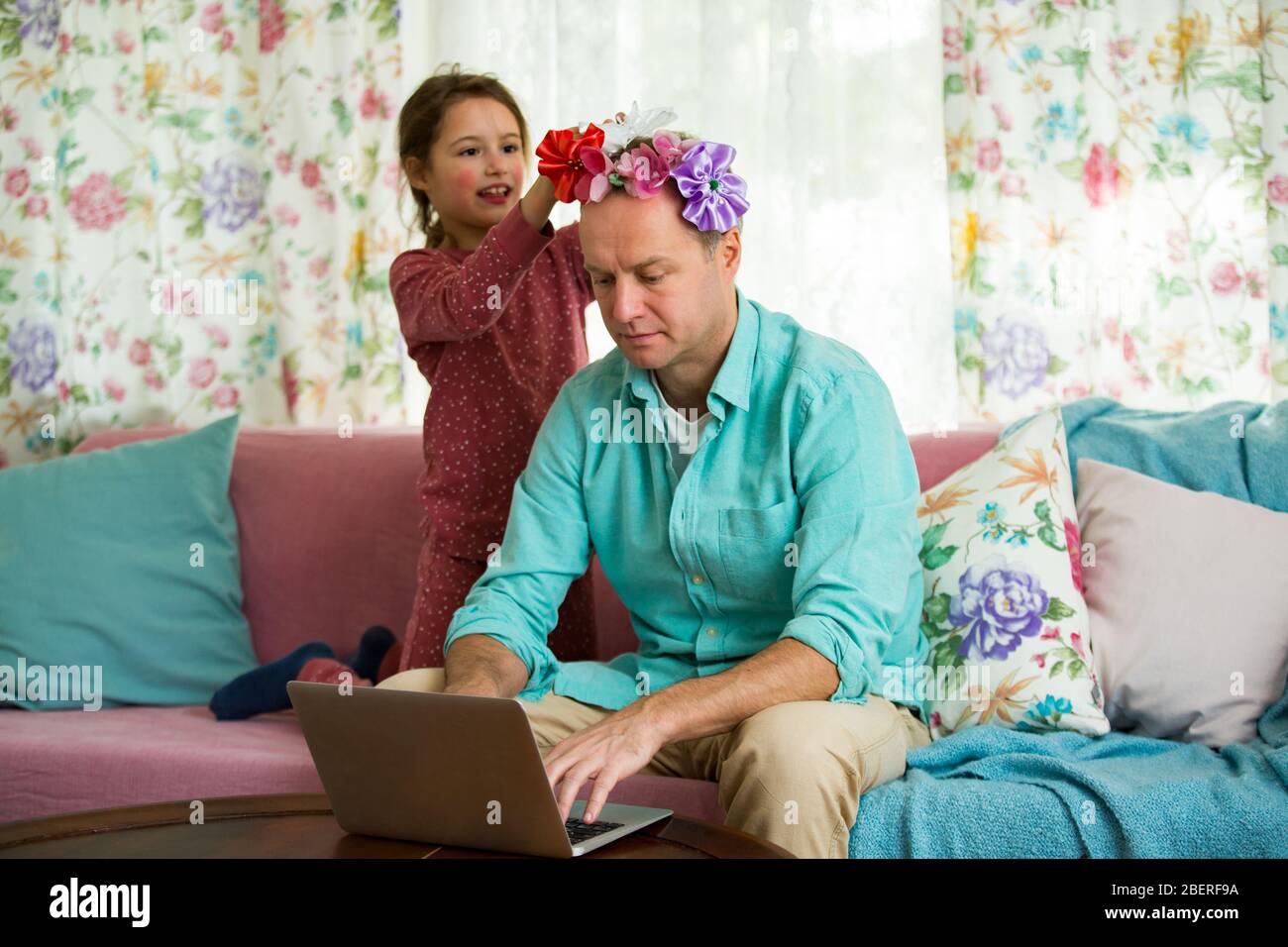 Child playing and disturbing father working remotely from home. Little girl combing daddy's hair and making hairstyle. Man on couch with laptop Stock Photo