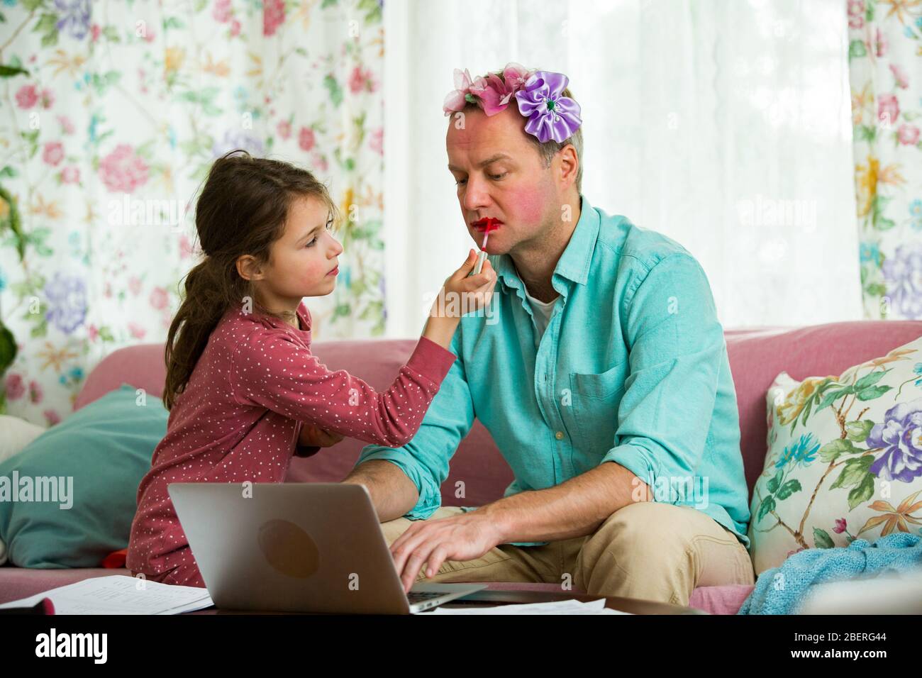 Child playing and disturbing father working remotely from home. Little girl applying makeup. Man sitting on couch with laptop. Family spending time to Stock Photo