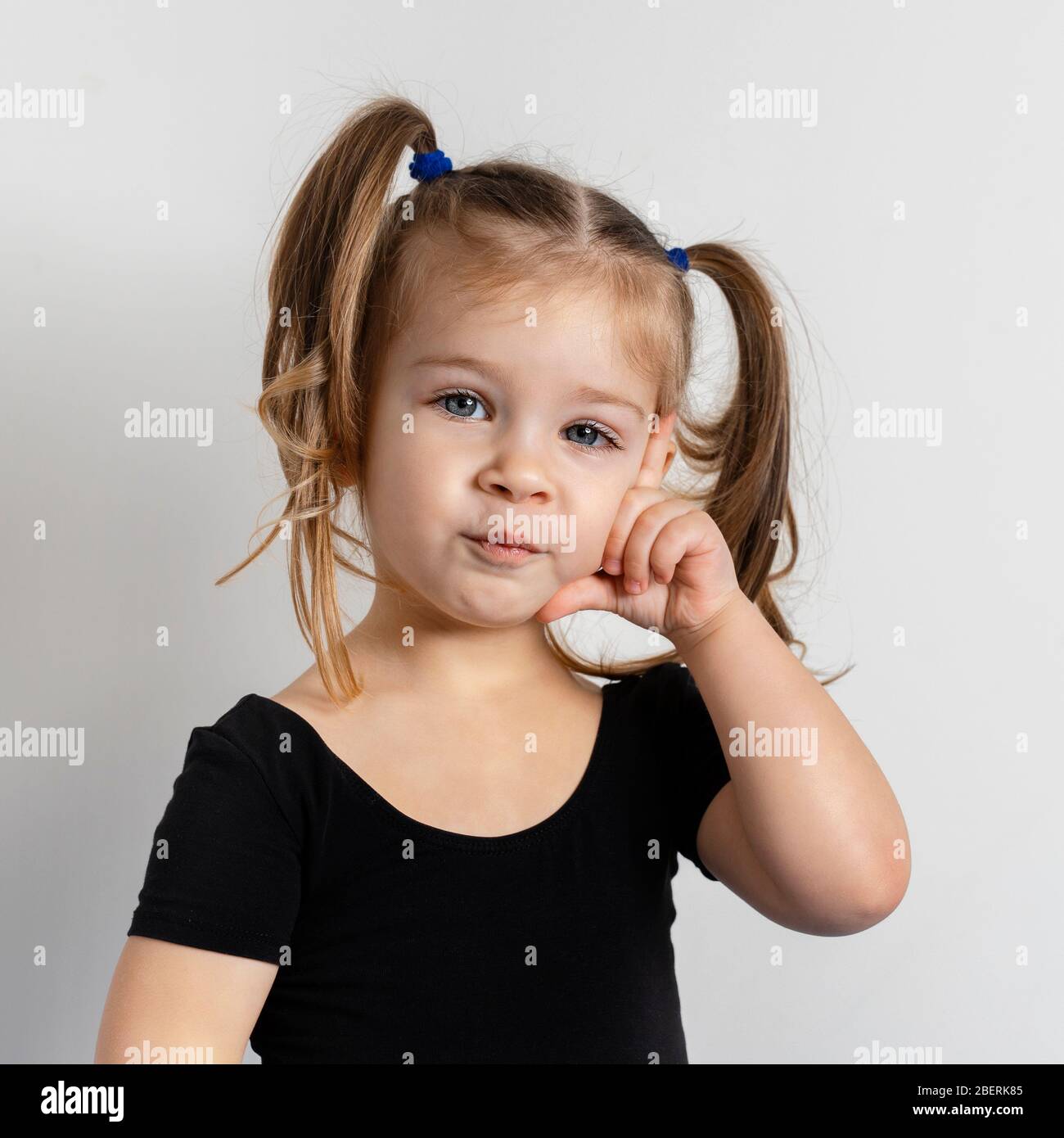 pretty little girl with two ponytails is standing in a pensive pose on a light background. Stock Photo