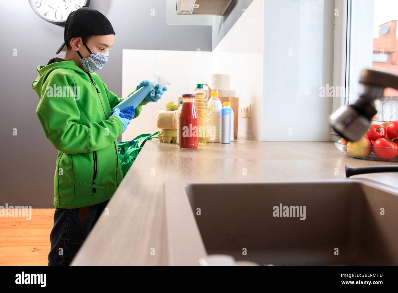 Coronavirus pandemic prevention. A boy wearing gloves and a mask uses a disinfectant liquid to wipe the food package upon delivery at home. Cleaning t Stock Photo