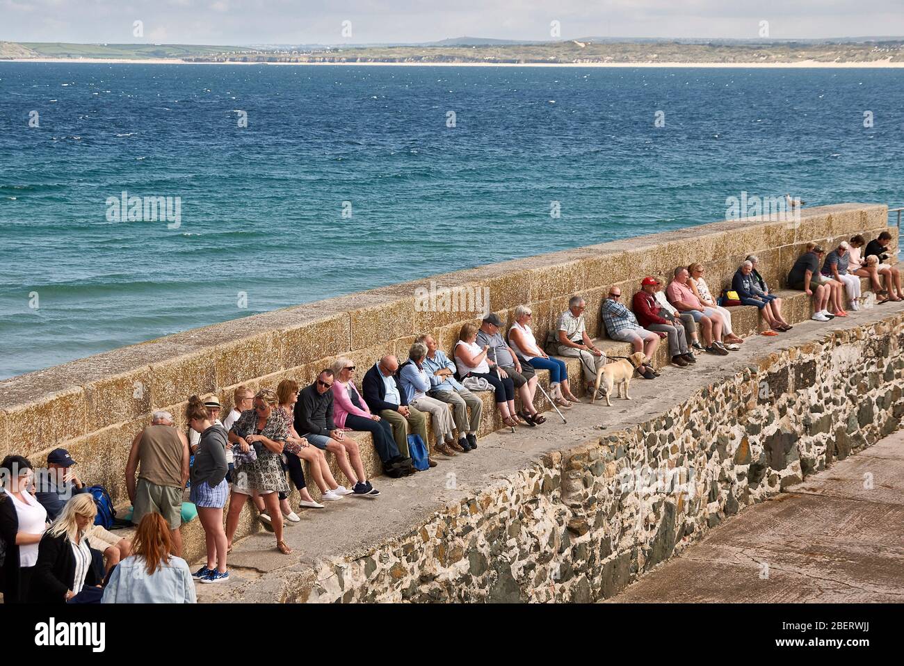 Visitors enjoy sitting in lee on the quay wall the view on the harbour of St Ives. Stock Photo