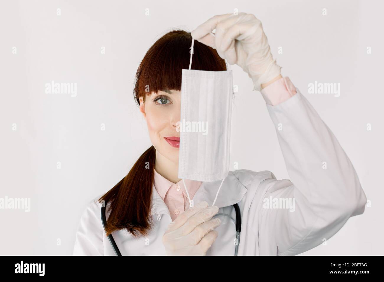 Young pretty dark haired woman doctor or nurse, wearing white coat, hides her face behind a medical mask, posing on isolated white background, copy Stock Photo