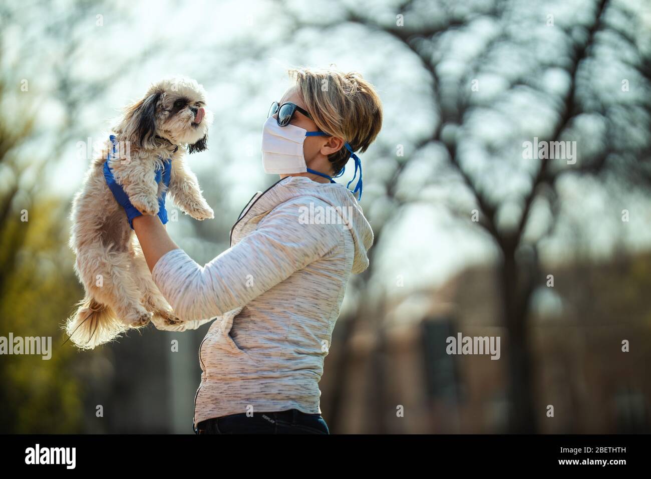 A woman in a medical protective mask is spending time with her dear cute little Shih Tzu dog on the city street path during flu virus outbreak and cor Stock Photo