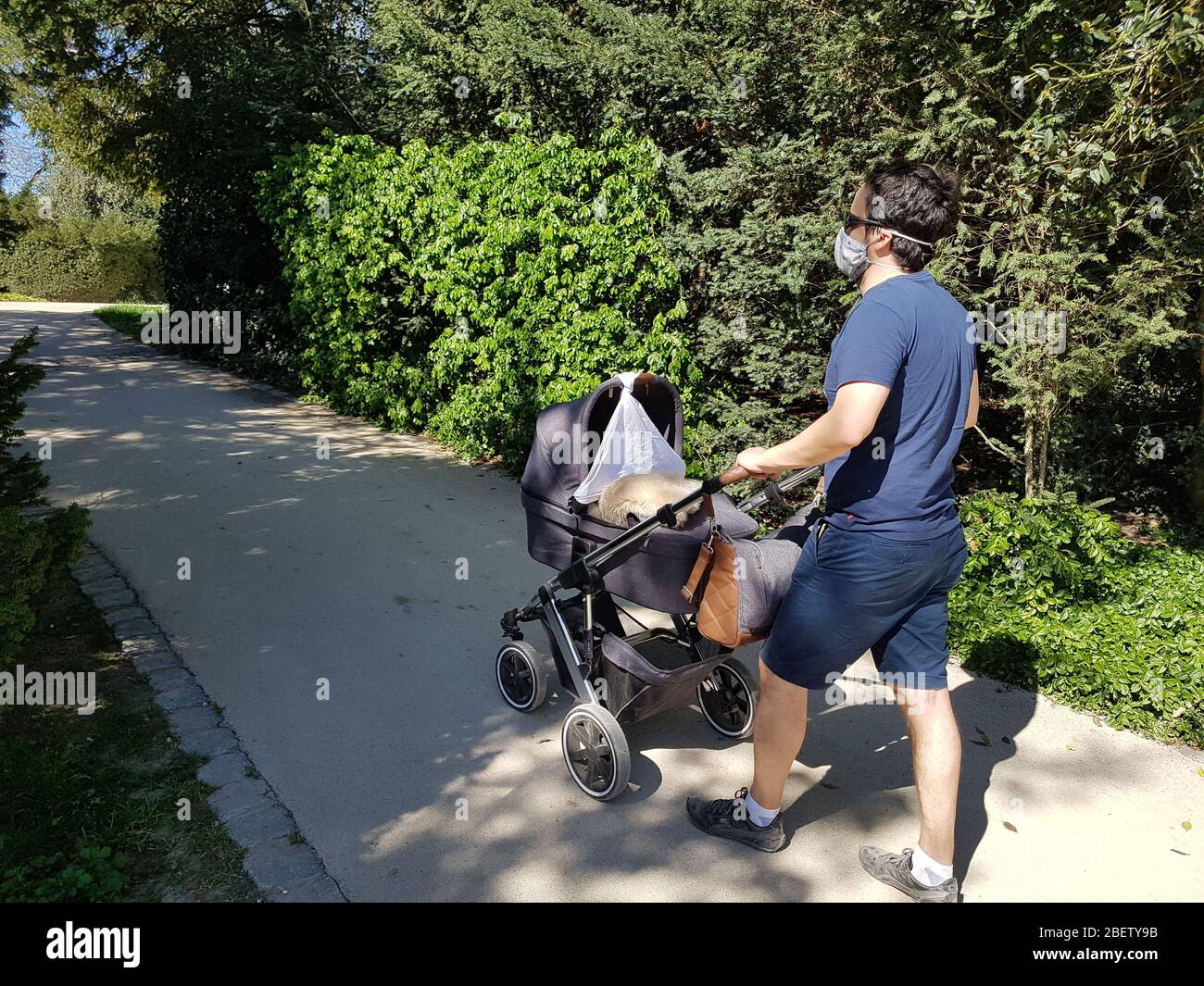 Person ( man / male father ) wear dust / protective / surgical / face mask while walking through park with baby buggy carriage - corona virus covid-19 Stock Photo