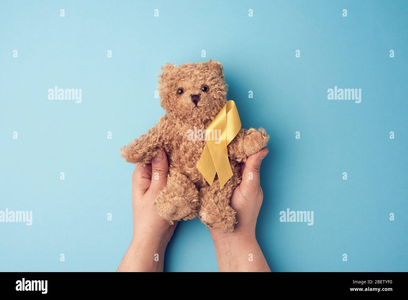 female hands hold a small teddy bear with a yellow ribbon folded in a loop on a blue background. concept of the fight against childhood cancer. proble Stock Photo