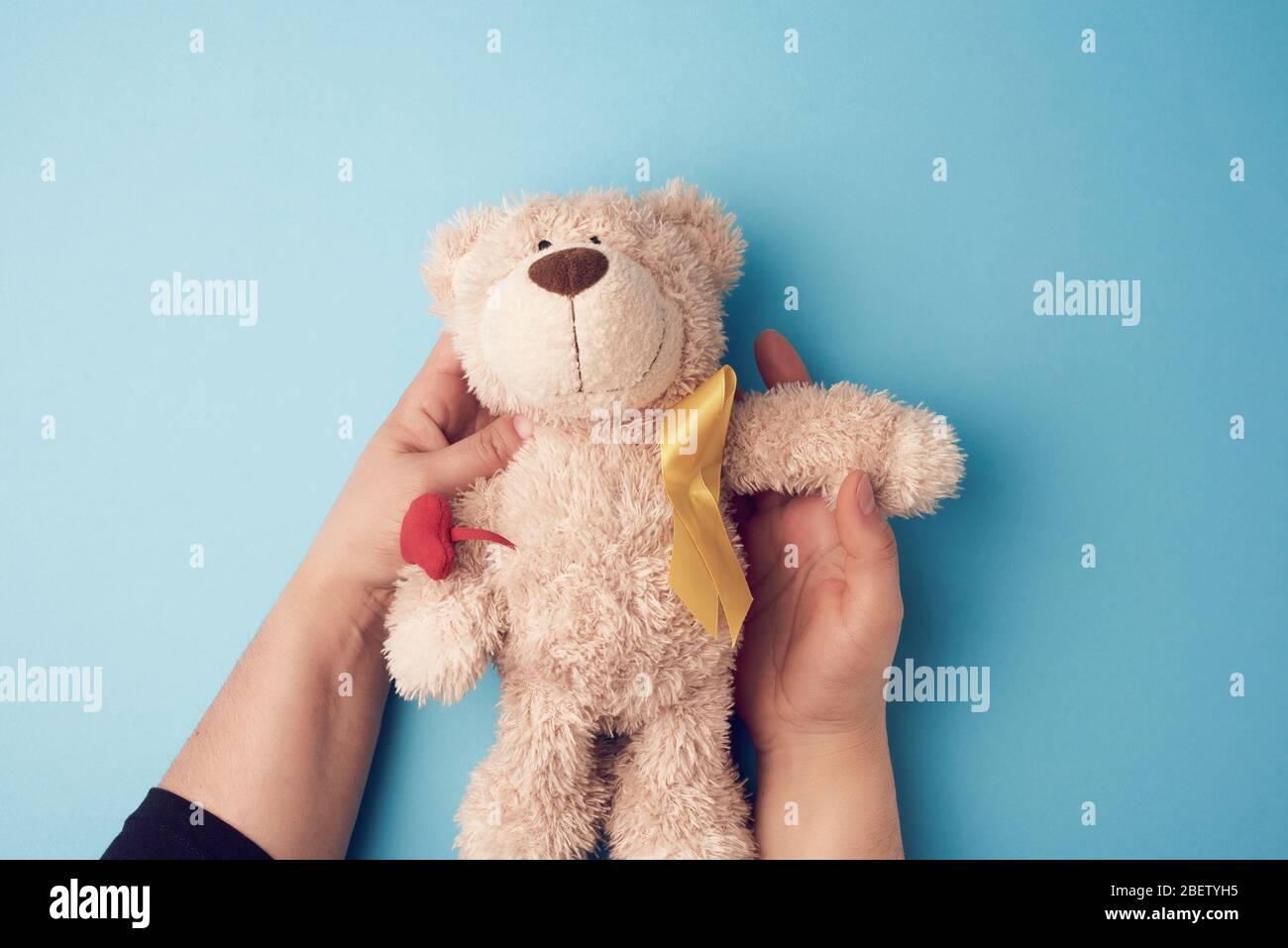 female hands hold a small teddy bear with a yellow ribbon folded in a loop on a blue background. concept of the fight against childhood cancer. proble Stock Photo