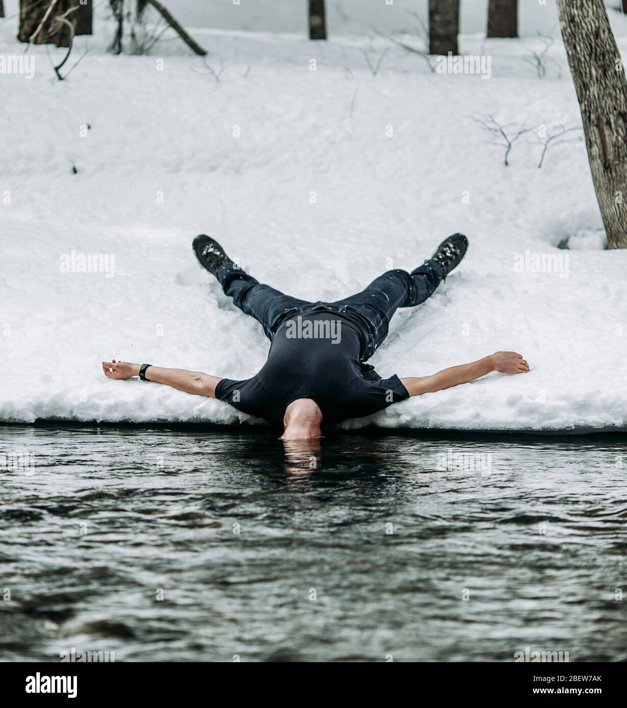 man lies on back in snow spread eagle with head underwater in river Stock Photo